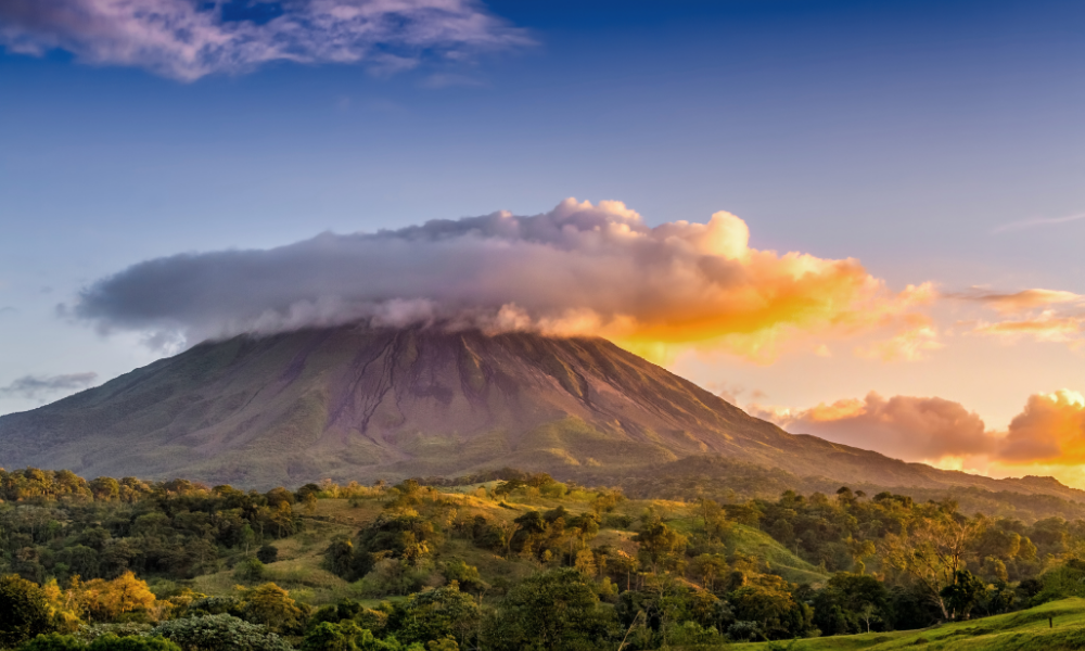 A stunning view of the Arenal Volcano with a dramatic cloud formation at sunset, surrounded by lush green vegetation.