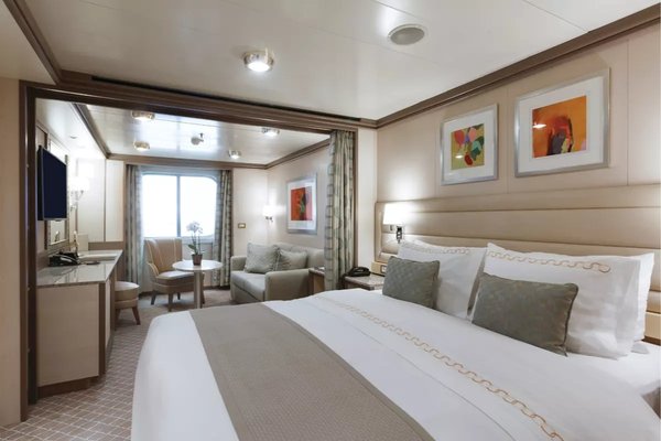 A spacious Vista Suite onboard a luxury cruise, featuring a large bed, elegant decor, a seating area, and a window offering natural light.